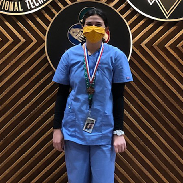 Medical Assisting - GNB Student standing in front of a wall with the GNBVT logo on it