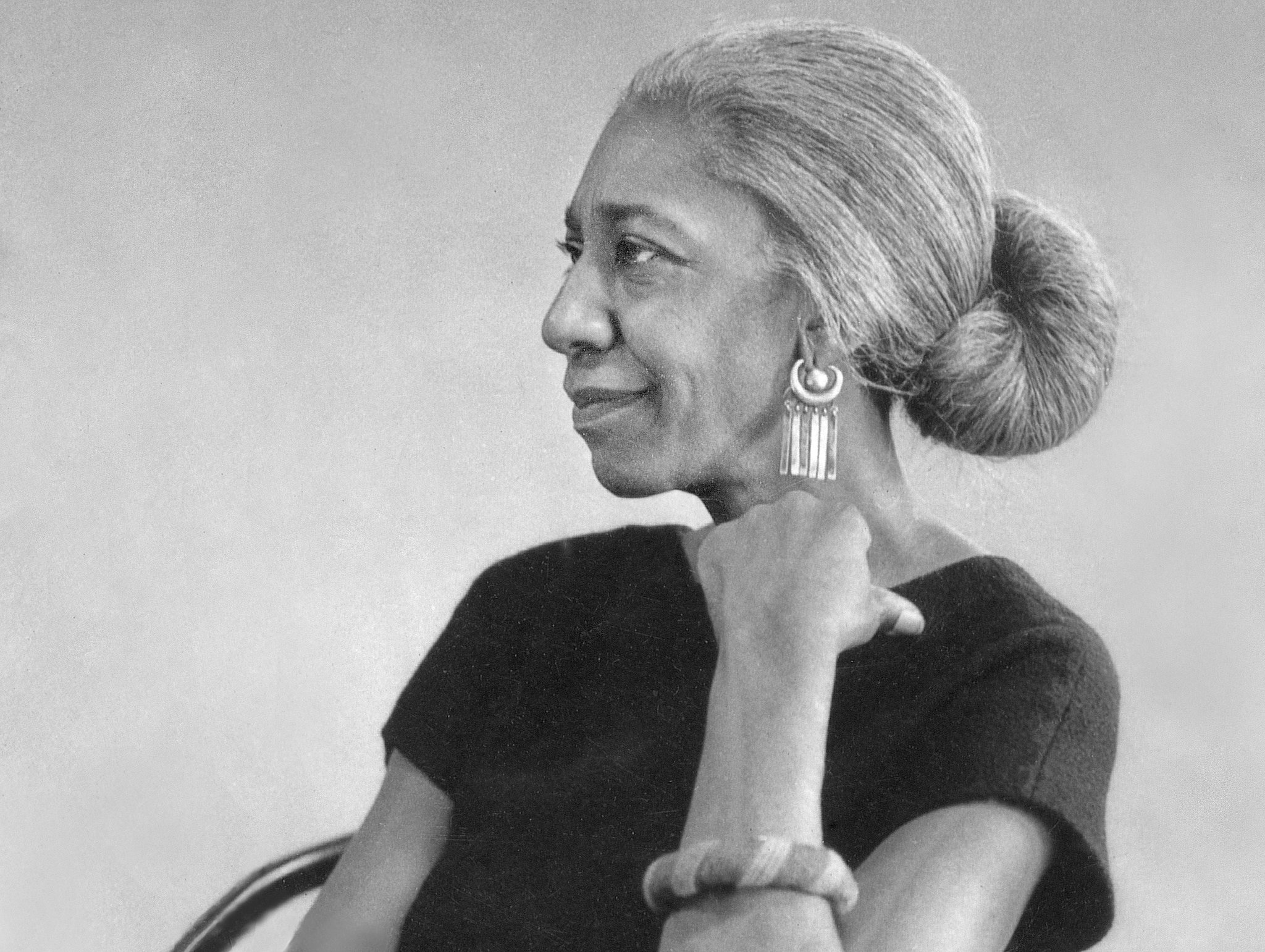 A black and white bust photo of Edna Lewis. She is sitting on a chair facing the left side of the photo. Her hand rests on her collar bone and she is wearing a black shirt, a bangle on her arm, and intricately designed earrings. Her hair is pulled back into a low bun.
