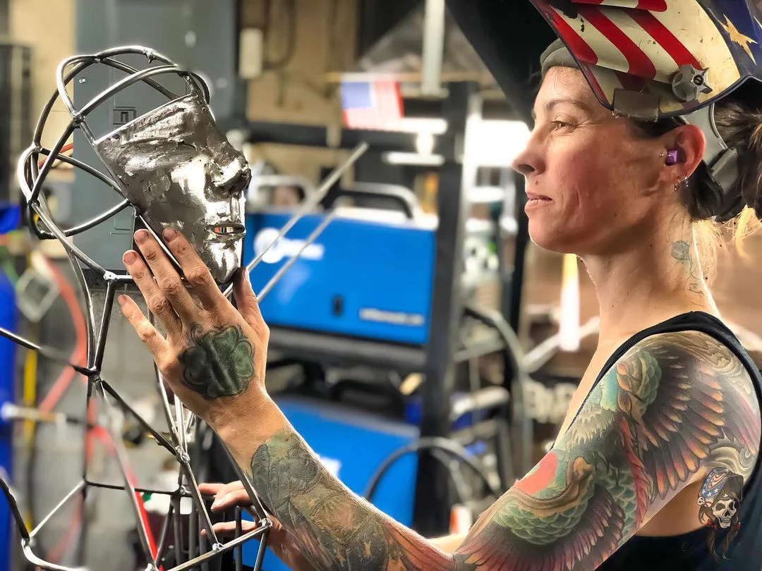 Image of Barbra Persons reaching her arm out to hold the face of an unfinished sculpture of a human. Persons is wearing her American flag welding helmet.