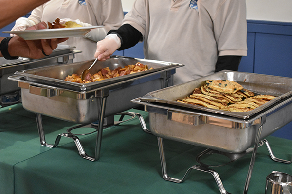 Bus Driver Appreciation 10 Two people serving potatoes and pancakes out of a metal heating tray.