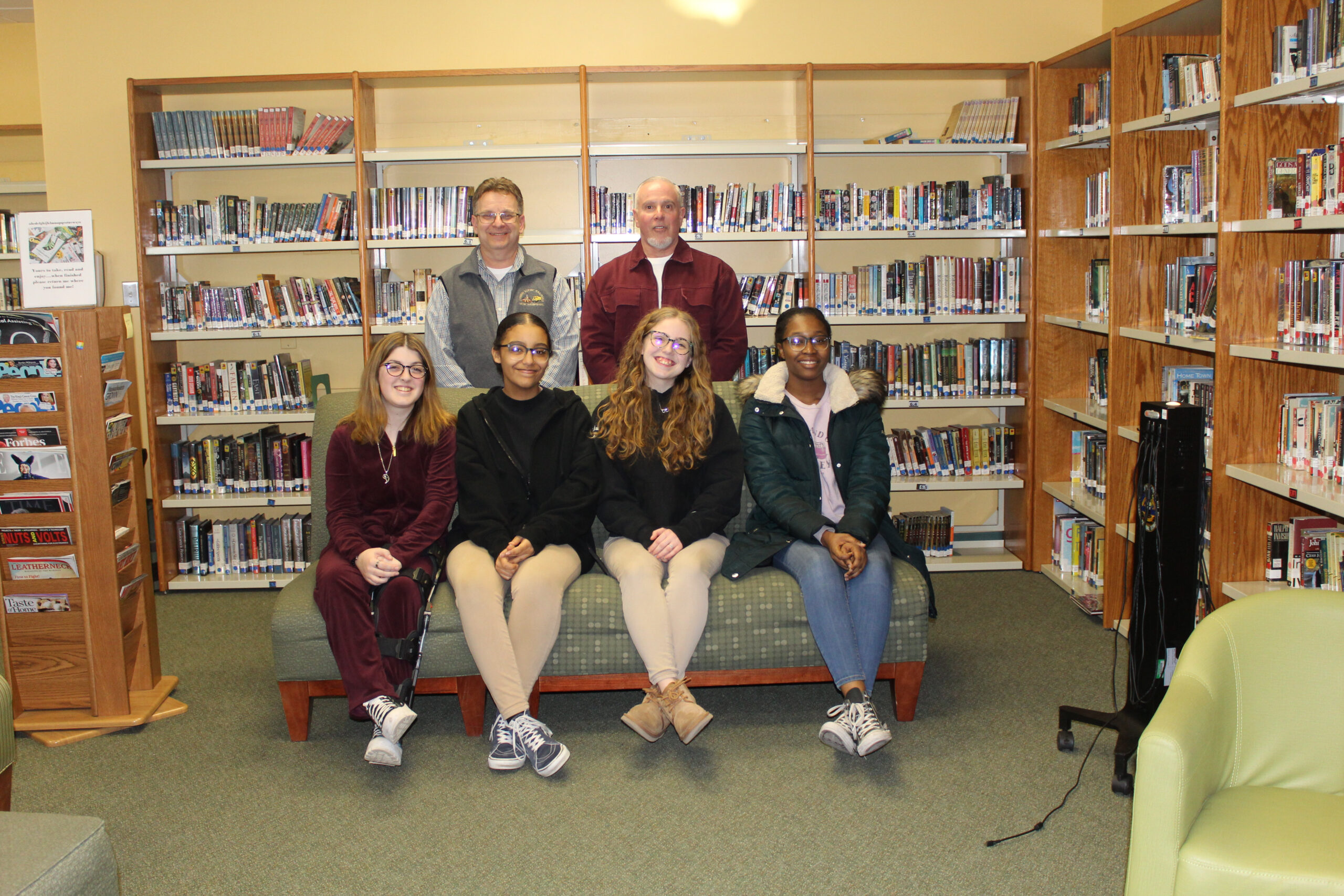 Photo of GNBVT's Book Club. With four students sitting on a couch and with advisors standing behind them in a library setting